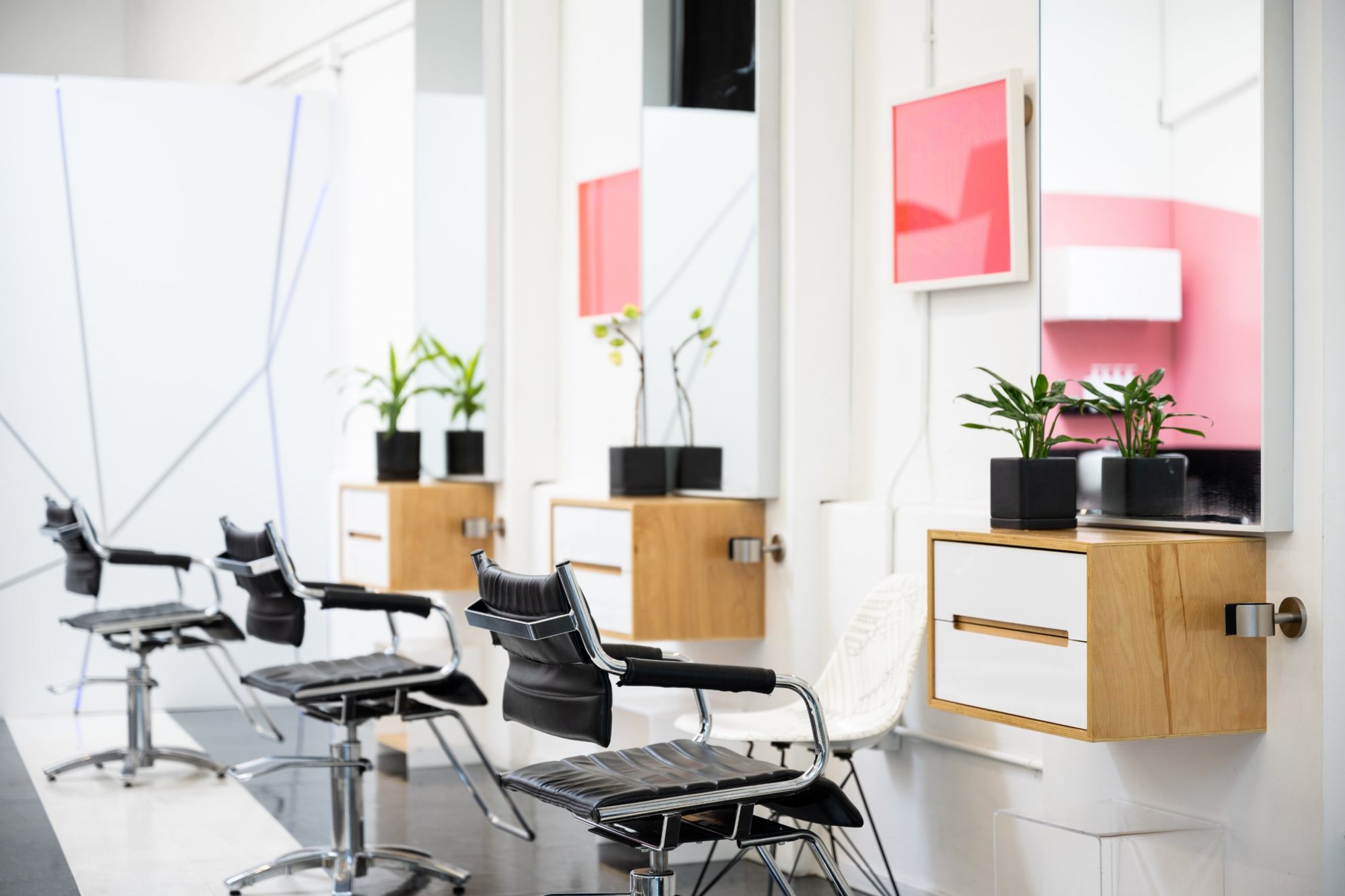 Three black styling chairs facing wall-mounted mirrors with plants, wooden cabinets and pink framed art at Cult in Portland